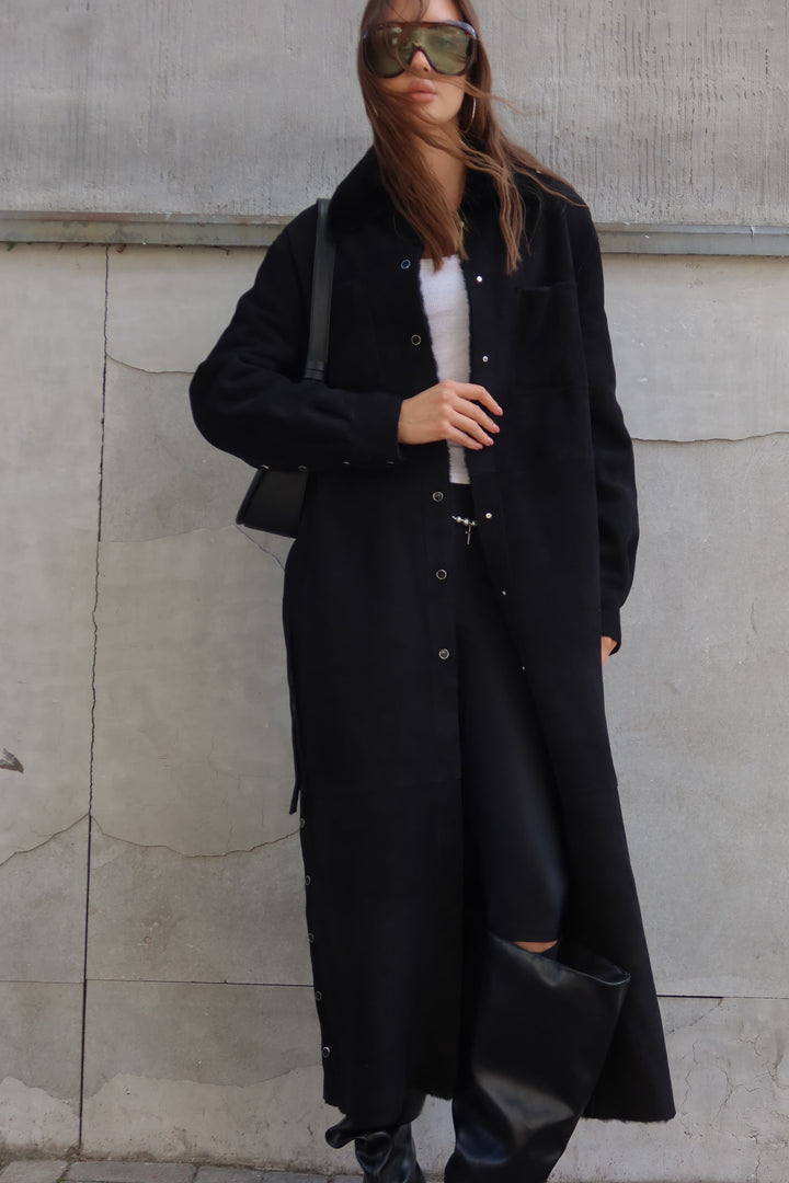 Elegant black merino sheepskin coat with long silhouette, belted waist, and button details for premium women's fashion.